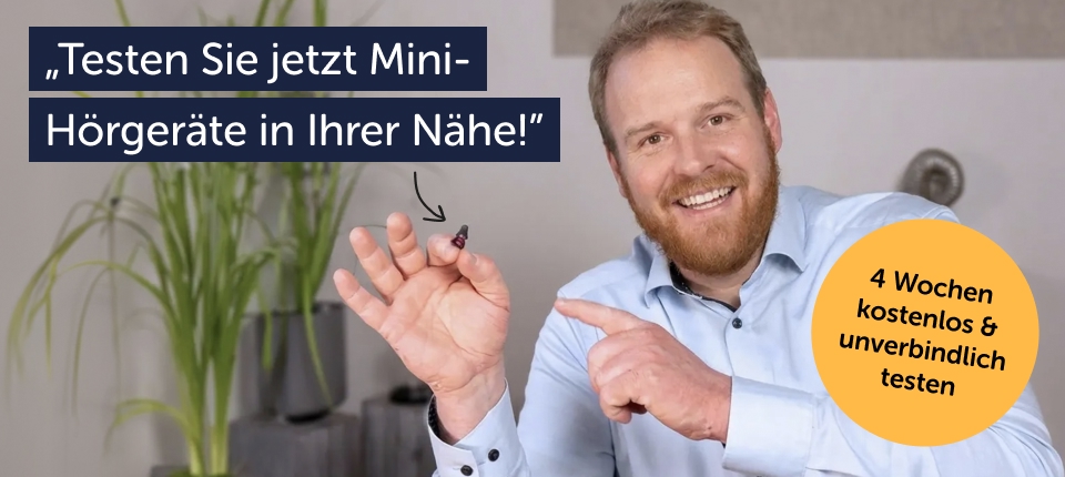 Ein lächelnder Mann mit Bart zeigt auf ein winziges, dunkelrotes Im-Ohr-Hörgerät, das er auf seiner Fingerkuppe hält.