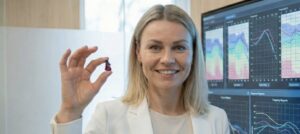 Eine lächelnde Expertin hält das winzige In-Ohr-Hörgerät audibene IX hoch; im Hintergrund zeigt ein Monitor audiologische Analysen zur Gehirnstimulation.