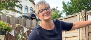 Melanie steht lächelnd in einem Garten und hält sich an einem Holztor fest. Im Hintergrund sind ein Haus, Pflanzen und ein Gartenzaun zu sehen.