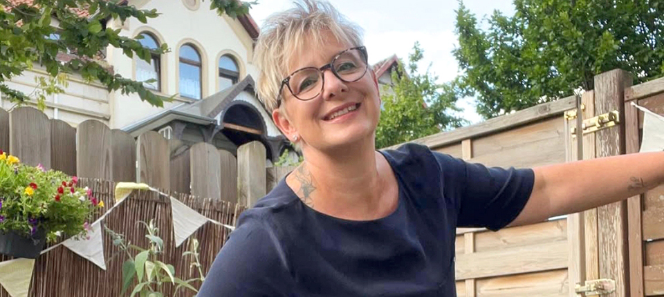 Melanie steht lächelnd in einem Garten und hält sich an einem Holztor fest. Im Hintergrund sind ein Haus, Pflanzen und ein Gartenzaun zu sehen.
