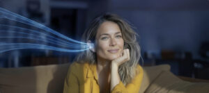 Eine Frau sitzt auf einem Sofa, während stilisierte blaue Schallwellen auf ihr Ohr zulaufen. Das Bild visualisiert das Thema Hörvermögen und Hör-Check.